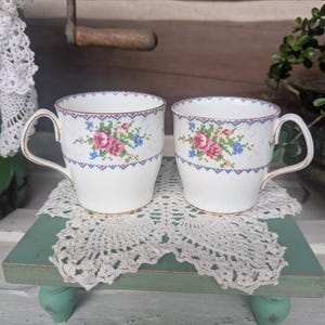 May include: Two white ceramic mugs featuring a delicate cross-stitch style floral pattern in pink, blue, and green. Each mug has a gold-trimmed rim and handle, with a repeating geometric border design encircling the upper portion of the body.