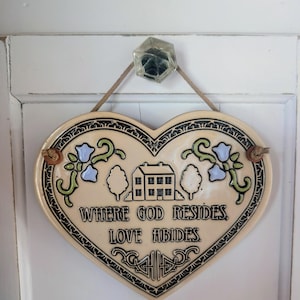 May include: A heart-shaped ceramic wall plaque with a cream-colored glaze. It features a central illustration of a house flanked by two trees, with blue flowers and green vines. The text reads: WHERE GOD RESIDES, LOVE ABIDES. It hangs from a brown cord.