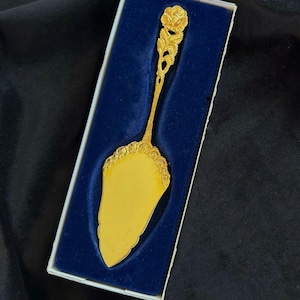 Könnte beinhalten: Ein goldfarbener Tortenheber mit floralem Design an Griff und Klingenrand, präsentiert in einer weißen Schachtel mit blauem Samtinnenraum. Der Heber hat eine dekorative Rose oben.