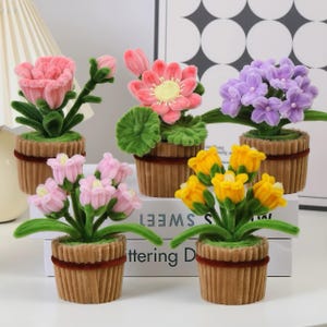 Puede incluir: Cinco arreglos florales artificiales en macetas pequeñas marrones con forma de magdalena. Las flores son rosas, moradas y amarillas, con tallos y hojas verdes. Las macetas tienen una banda decorativa marrón. Sobre una superficie blanca.