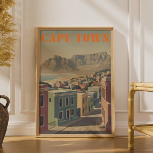 Può includere: Una stampa artistica incorniciata con un'illustrazione stilizzata di Città del Capo e la scritta CAPE TOWN in arancione in alto. L'immagine raffigura una strada di una città costiera che conduce verso una grande catena montuosa.