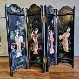 Puede incluir: Un biombo plegable negro de cuatro paneles con figuras en relieve vestidas con atuendos tradicionales. Cada panel muestra elementos paisajísticos pintados, incluyendo árboles y una luna creciente, con detalles dorados y bisagras de metal.