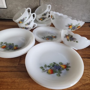 Op de afbeelding: Een set wit melkglas servies met een fruitpatroon van peren en blauwe druiven. De collectie bestaat uit gestapelde dinerborden, saladeborden, een grote kom met geschulpte rand, een kleinere kom en diverse theekopjes.