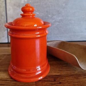 Pode incluir: Um recipiente de cerâmica laranja brilhante com tampa a condizer e um topo arredondado. O recipiente cilíndrico tem um acabamento liso e brilhante, com uma borda alargada e uma base elevada. Ao lado, encontra-se uma pequena pá de madeira.
