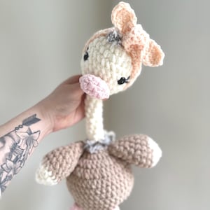 May include: A handmade crochet giraffe toy in cream, tan, and pink. The giraffe has a long neck, small black eyes, and a pink muzzle. The ears and legs are pink, and the body is tan. The toy is soft and plush.