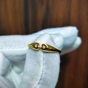 Op de afbeelding: Een gouden ring met twee hartvormige segmenten die met elkaar verbonden zijn. Het linkerhart is gegraveerd met de letter E en het rechterhart is gegraveerd met de letter B. Een kleine, heldere, ronde edelsteen is tussen de twee harten gezet.