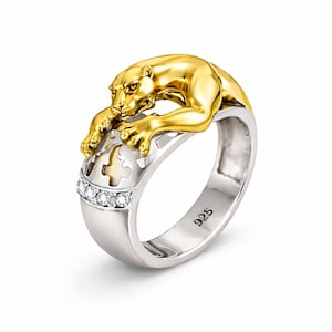 Puede incluir: Anillo bicolor con una pantera dorada sobre una banda plateada. La pantera presenta detalles de pelaje texturizado y una expresión concentrada. La banda tiene un patrón decorativo y una fila de pequeñas piedras claras.