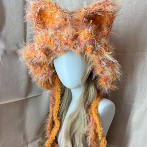 May include: A whimsical, handmade orange and yellow fox ear hat. The hat is made of fluffy, textured yarn with colorful flecks. Long, orange yarn strands hang down the sides. The hat is displayed on a mannequin head with long, blonde hair.