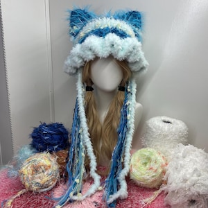 Op de afbeelding: Een blauw-witte imitatiebont hoed met kattenoren, gedragen door een mannequin met blonde vlechten. De hoed heeft een zachte textuur en is omgeven door kleurrijke bollen garen in blauwe, witte en gele tinten. Het oorontwerp van de hoed voegt een speels tintje toe.