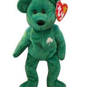 May include: A green plush bear with a white shamrock on its chest and black eyes. The bear has a red heart-shaped tag attached to its ear. The bear is standing upright against a white background.
