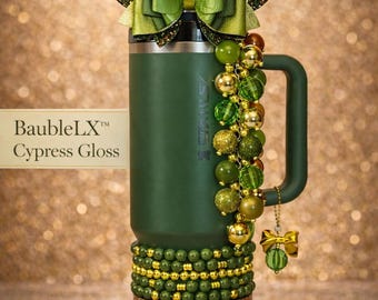 Op de afbeelding: Een matte bosgroene beker met handvat, versierd met een grote groene strik, een kralenbedel met groene en gouden bollen, en een basis omwikkeld met een gestructureerde gouden band. De tekst BaubleLX Cypress Gloss is zichtbaar op een label.