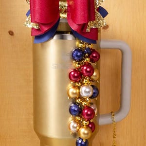 Op de afbeelding: Een goudkleurige geïsoleerde beker met een grijze handgreep, versierd met een rode en gouden strik. Een kralenbedel met rode, gouden, blauwe en witte kralen hangt aan de handgreep. De beker is versierd met een cluster van kralen.