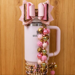 May include: A light pink insulated tumbler featuring a large pink and gold glitter bow on top. The tumbler is decorated with a beaded charm strand of pink, gold, and leopard print beads, a beaded base wrap, and a dark brown rhinestone bottom band.