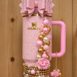 May include: A pink Stanley tumbler featuring a floral pattern, a large glittery pink bow on top, and a handle. The cup is decorated with a beaded charm strand, a pink rose accent, and multiple rows of gold and white beads around the base.