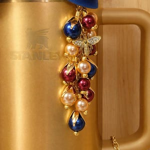 Könnte beinhalten: Ein goldfarbener Stanley-Becher mit einem dekorativen Anhänger. Der Anhänger zeigt einen Schmuck-Bienenanhänger und Cluster aus perlenartigen Perlen in Blau-, Rot- und Cremetönen. Oben am Becher ist ein blaues und rotes Band gebunden.