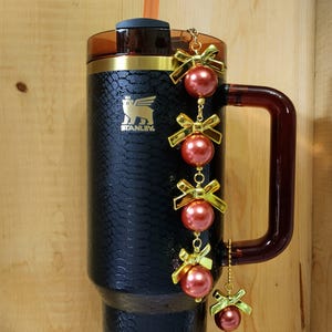 May include: A black Stanley tumbler with a textured pattern, gold accents, and a brown handle. The tumbler is decorated with a chain of red ornaments and gold bows. A brown straw is inserted into the lid.