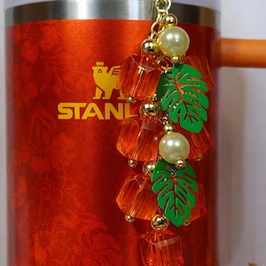 May include: A red Stanley tumbler with a gold logo and an orange handle. A decorative charm hangs from the handle, featuring orange crystal beads, green leaf charms, gold beads, and faux pearl accents.