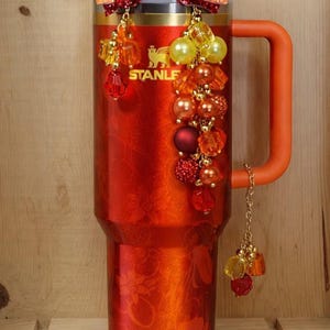 May include: A red Stanley tumbler with an orange handle and decorative beaded charms. The tumbler has the brand name "STANLE" printed on it. The charms are in shades of red, orange, and yellow.