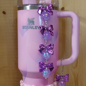 May include: A pink Stanley tumbler with a handle, decorated with purple bows and iridescent orbs. The base is adorned with pink roses and pearls. A clear straw is inserted into the lid.