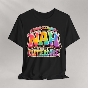 May include: Black t-shirt with a colorful graphic that reads "SPRING CLEANING? NAH THIS IS CLUTTERCORE." The text is in a bubble letter style with a rainbow gradient. The shirt is a crew neck style.