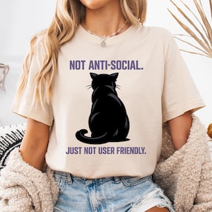 Peut inclure: T-shirt beige avec le texte "NOT ANTI-SOCIAL. JUST NOT USER FRIENDLY." et une silhouette de chat noir. Le chat est de dos. Le texte est en violet.