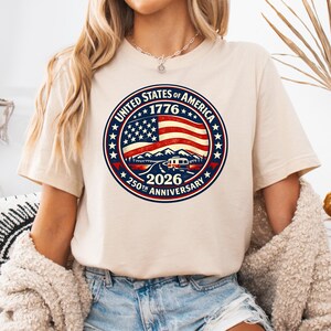 Pode incluir: T-shirt bege com um design circular que comemora o 250º aniversário dos Estados Unidos da América em 2026. O design inclui a bandeira americana, montanhas, uma caravana e o texto "United States of America 1776 250th Anniversary 2026".