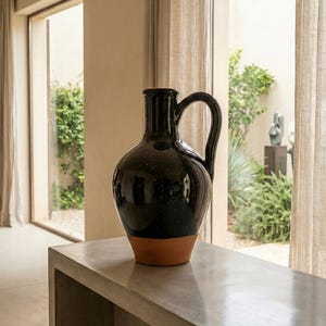 Puede incluir: Una jarra de cerámica negra con acabado moteado y base color terracota. La jarra tiene un asa curva y está sobre una superficie de hormigón. El fondo incluye una ventana con cortinas transparentes y vegetación.
