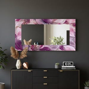 Puede incluir: Un espejo de pared rectangular con un borde decorativo con un diseño de hojas en tonos morados y rosas. El espejo está montado en una pared gris oscuro sobre un mueble negro. Un reloj digital muestra la hora.