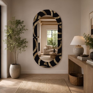 May include: An oval wall mirror featuring a wide frame with a black and gold abstract curved pattern. The reflective surface shows a room interior with a sofa, fireplace, and wooden furniture. The mirror is mounted on a plain light-coloured wall.