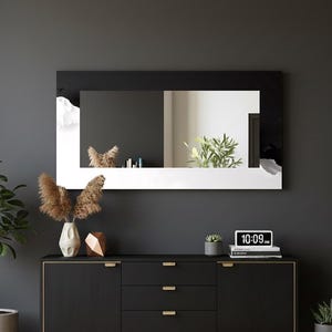 Puede incluir: Un espejo rectangular con marco blanco y negro cuelga sobre un mueble negro. El espejo refleja una habitación con plantas. Un reloj digital muestra las 10:09 AM. Los elementos decorativos incluyen un jarrón con flores secas y una escultura geométrica.