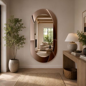 May include: A large, oval-shaped wall mirror featuring a wide, reflective copper-toned frame with a smooth, curved surface. The mirror is mounted vertically on a wall, displaying a clear reflection of a living room interior with a sofa and fireplace.