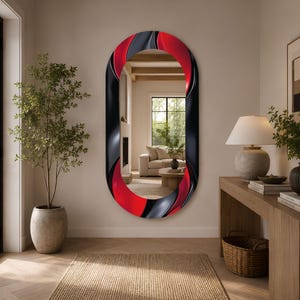 May include: An oval wall mirror featuring a wide, decorative frame with a fluid, abstract pattern in bold red and black. The reflective surface shows a clear view of a living room interior, including a sofa, a fireplace, and a wooden coffee table.