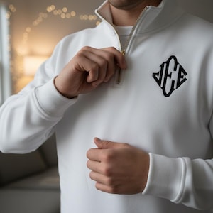 Puede incluir: Sudadera blanca con cremallera dorada y logotipo de monograma negro. La sudadera tiene cuello y puños acanalados. La persona se está subiendo la cremallera de la sudadera.