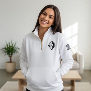 Könnte beinhalten: Weißes Sweatshirt mit Stehkragen, goldenem Reißverschluss, Vordertasche und schwarzem gesticktem Logo. Das Sweatshirt hat auch einen kleinen amerikanischen Flaggenaufnäher am Ärmel. Die Person trägt das Sweatshirt und lächelt.