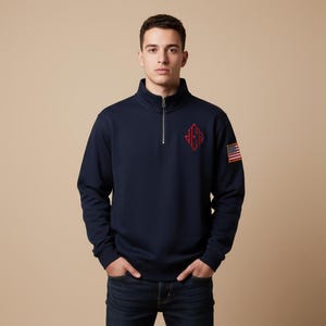 Puede incluir: Sudadera azul marino con cuello con cremallera. Cuenta con un emblema rojo bordado en el pecho y un pequeño parche de la bandera estadounidense en la manga. La sudadera está hecha de un material suave y cómodo.