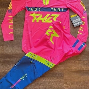 Puede incluir: Un conjunto de camiseta y pantalón de motocross rosa y azul. La camiseta y el pantalón presentan la marca "THOR" en amarillo y azul. La camiseta tiene mangas largas y el pantalón es azul con detalles rosas. Incluye una etiqueta.