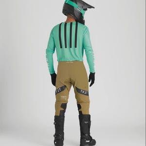 Może przedstawiać: Strój do jazdy na motocyklu crossowym, składający się z turkusowej koszulki z długim rękawem z trzema czarnymi paskami, spodni w kolorze khaki, czarnych butów i czarnego kasku z turkusowymi detalami. Strój jest przeznaczony do jazdy na motocyklu crossowym.