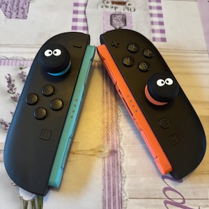 Puede incluir: Dos mandos Joy-Con negros para Nintendo Switch, uno turquesa y otro naranja, con agarres negros para el pulgar con ojos blancos. Los mandos tienen botones y crucetas negros. Fondo claro con patrones.
