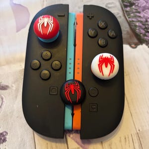 May include: Black Nintendo Switch Joy-Con controllers with red, white, and black Spider-Man themed thumb grips. The controllers are split with blue and orange accent strips.