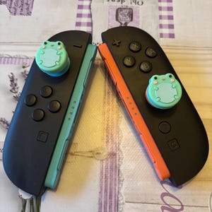 May include: Two black Nintendo Switch Joy-Con controllers with teal and orange sides. Each controller has a light green frog thumb grip. The controllers are on a patterned surface. The controllers are for a games console.