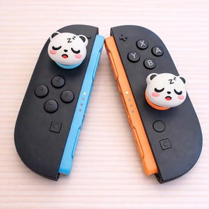 Op de afbeelding: Twee zwarte gamecontrollers met blauwe en oranje accenten, elk met een slapend berenontwerp. De controllers hebben knoppen en richtingspads. De tekst bovenaan luidt "Switch2 / PS5 / Xbox".