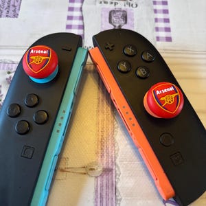 Könnte beinhalten: Zwei schwarze Gamecontroller mit dem Arsenal-Logo auf den Joystick-Tasten. Einer hat eine türkisfarbene Seite, der andere eine orangefarbene Seite. Die Controller liegen auf einer hellen Oberfläche.
