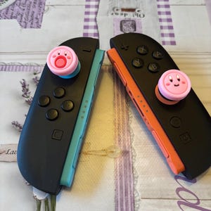 May include: Two black Nintendo Switch Joy-Con controllers with pink and blue accents. Each controller has a pink thumb grip featuring a cartoon character. One controller has a blue side, and the other has an orange side.