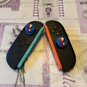 May include: Two black Nintendo Switch Joy-Cons with Deadpool-themed thumb grips. One Joy-Con has a teal side, the other an orange side. The thumb grips are blue with a Deadpool mask design.