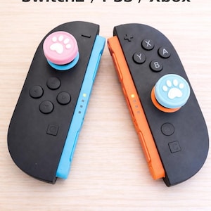 Puede incluir: Dos mandos de juego negros con detalles azules y naranjas. Cada mando tiene una empuñadura para el pulgar con huella de pata, una rosa y otra azul. El texto "Switch2 / PS5 / Xbox" está encima de los mandos.