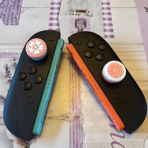 May include: Two black Nintendo Switch Joy-Con controllers. One has a light blue side and the other has an orange side. Both have a white and pink flower design on the joystick. The controllers are resting on a patterned surface.