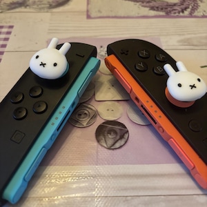 May include: Two black Nintendo Switch Joy-Con controllers, one with a teal side and the other with an orange side. Each controller has a white bunny-shaped thumb grip. The controllers are on a patterned surface.