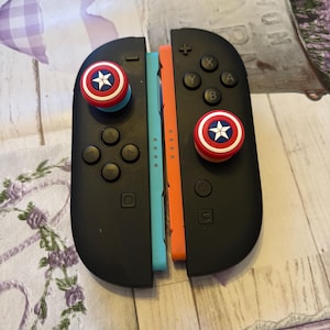 Peut inclure: Manettes Joy-Con noires pour Nintendo Switch, avec côtés bleus et oranges. Les manettes ont des boutons noirs et des protège-pouces en forme de bouclier Captain America. Sur une surface à motifs.