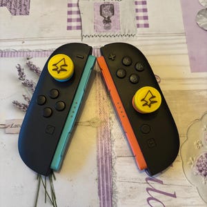 May include: Two black game controllers with blue and orange accents. Each controller has a yellow button with a tree-like design. The controllers are lying on a patterned surface with the word "Lav" visible.