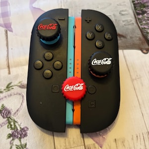 May include: Black Nintendo Switch Joy-Con controllers customized with Coca-Cola bottle cap designs. The controllers have black buttons and a red, white, and blue color scheme. The Coca-Cola logo is visible on the caps.
